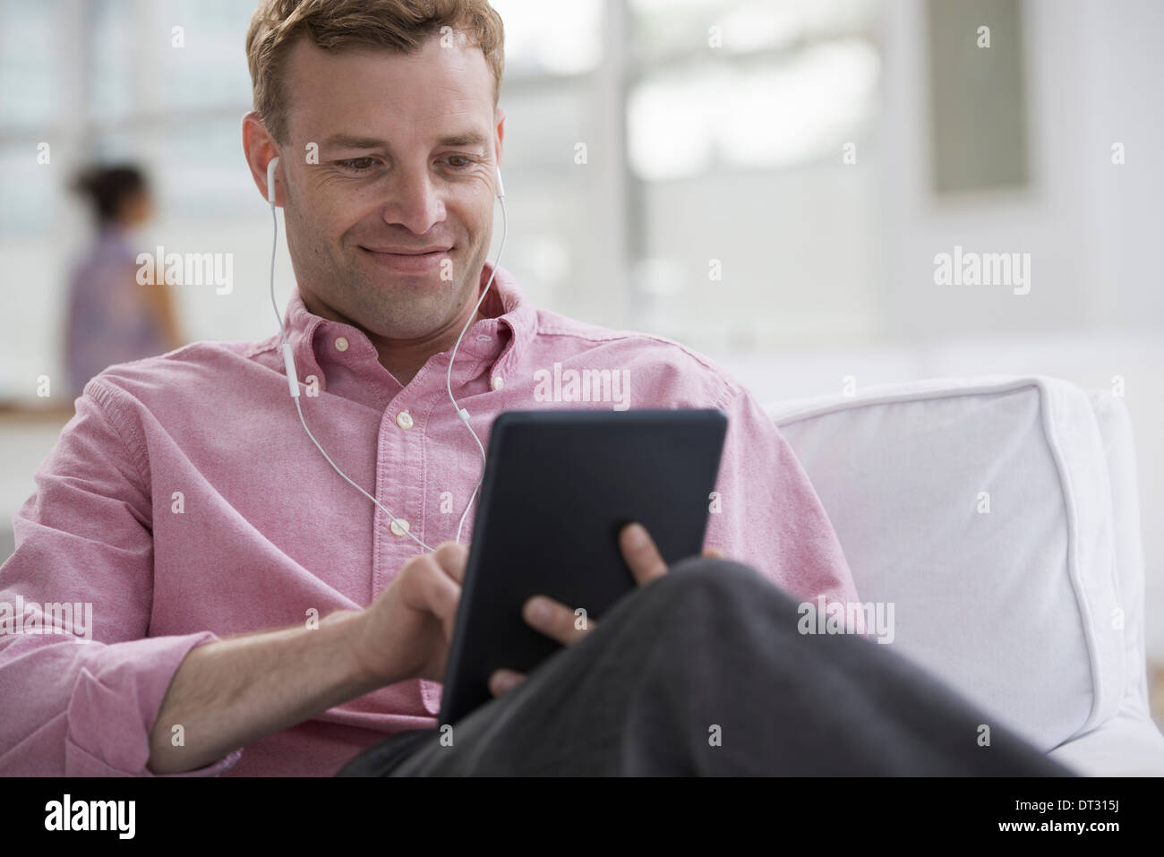 A man in a pink shirt sitting smiling using a digital tablet Wearing earphones Stock Photo