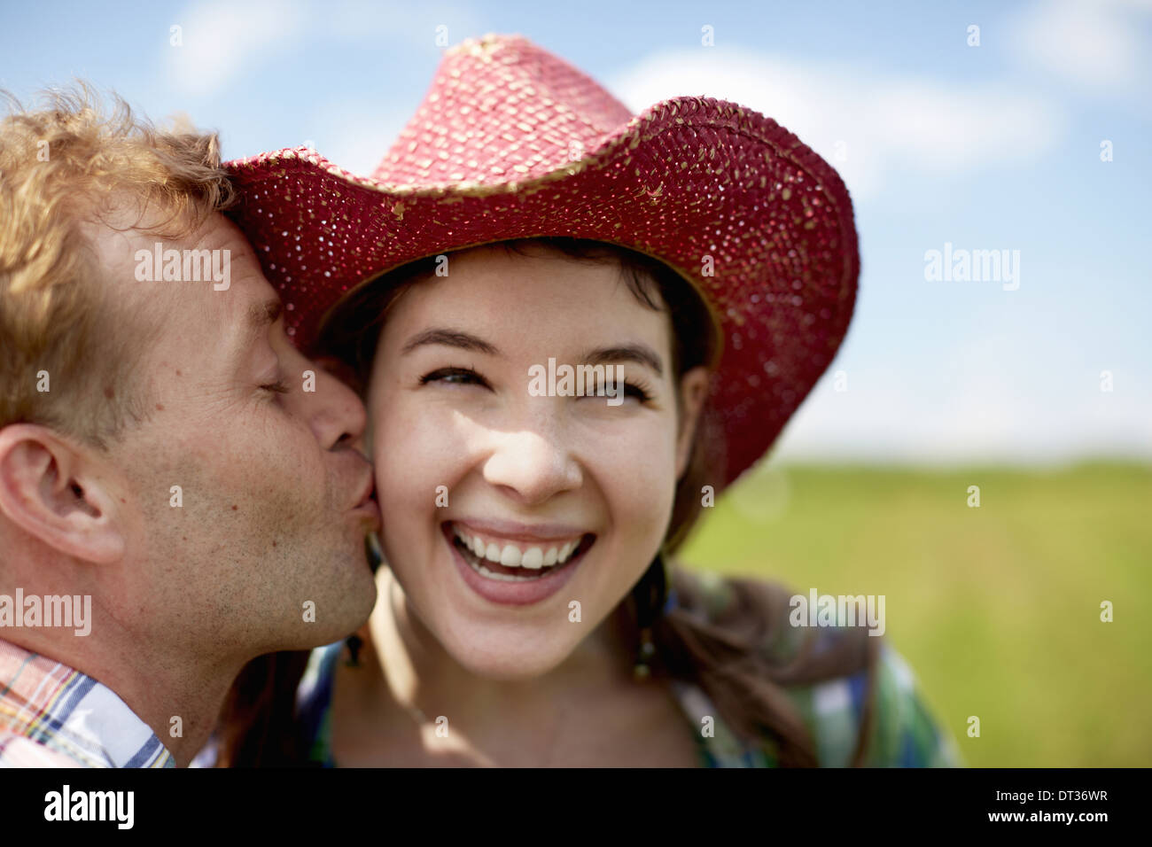 A young woman in a wide-brimmed pink hat being kissed on the cheek by a young man Stock Photo