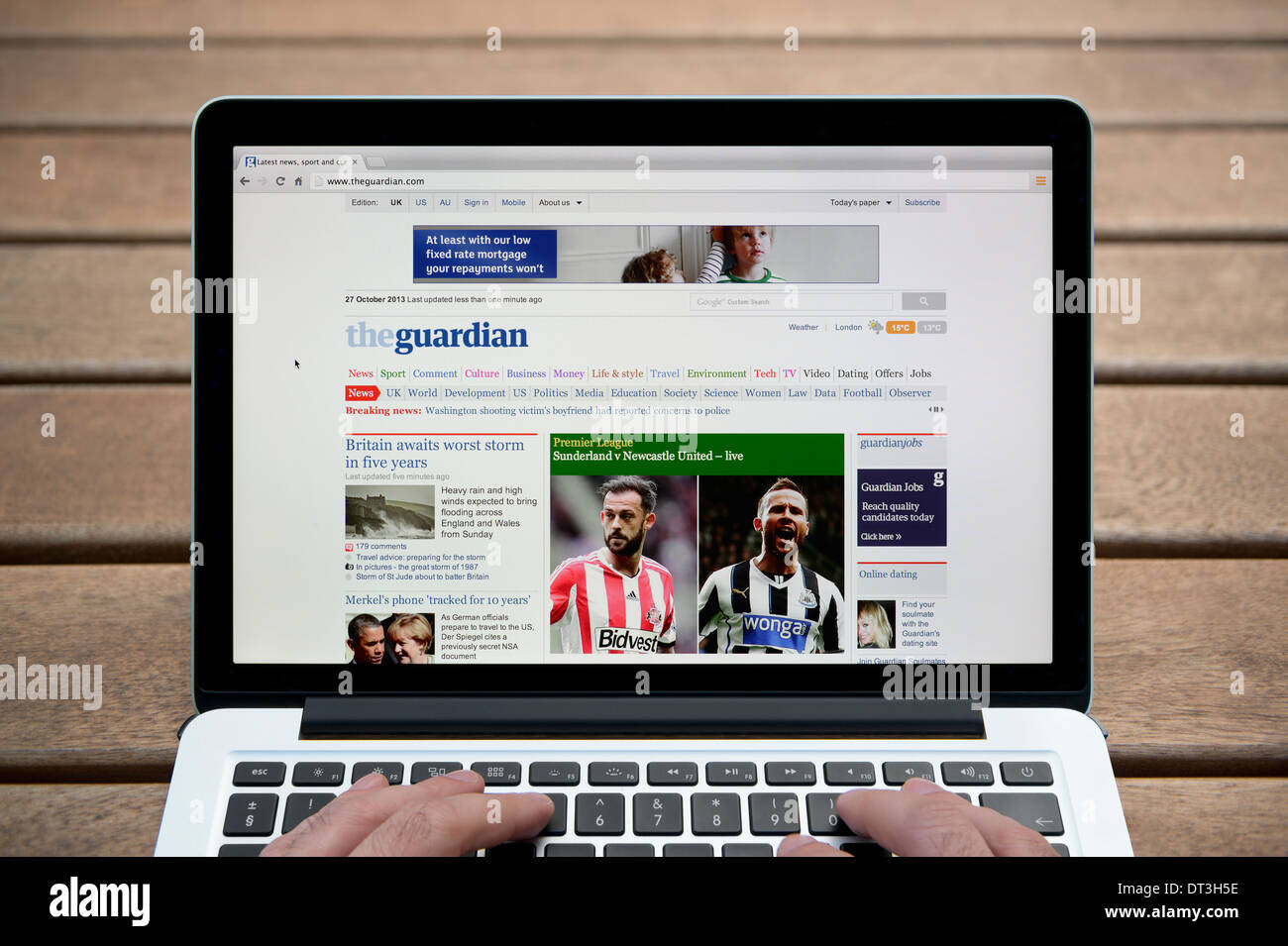 The Guardian website on a MacBook against a wooden bench outdoor background including a man's fingers (Editorial use only). Stock Photo
