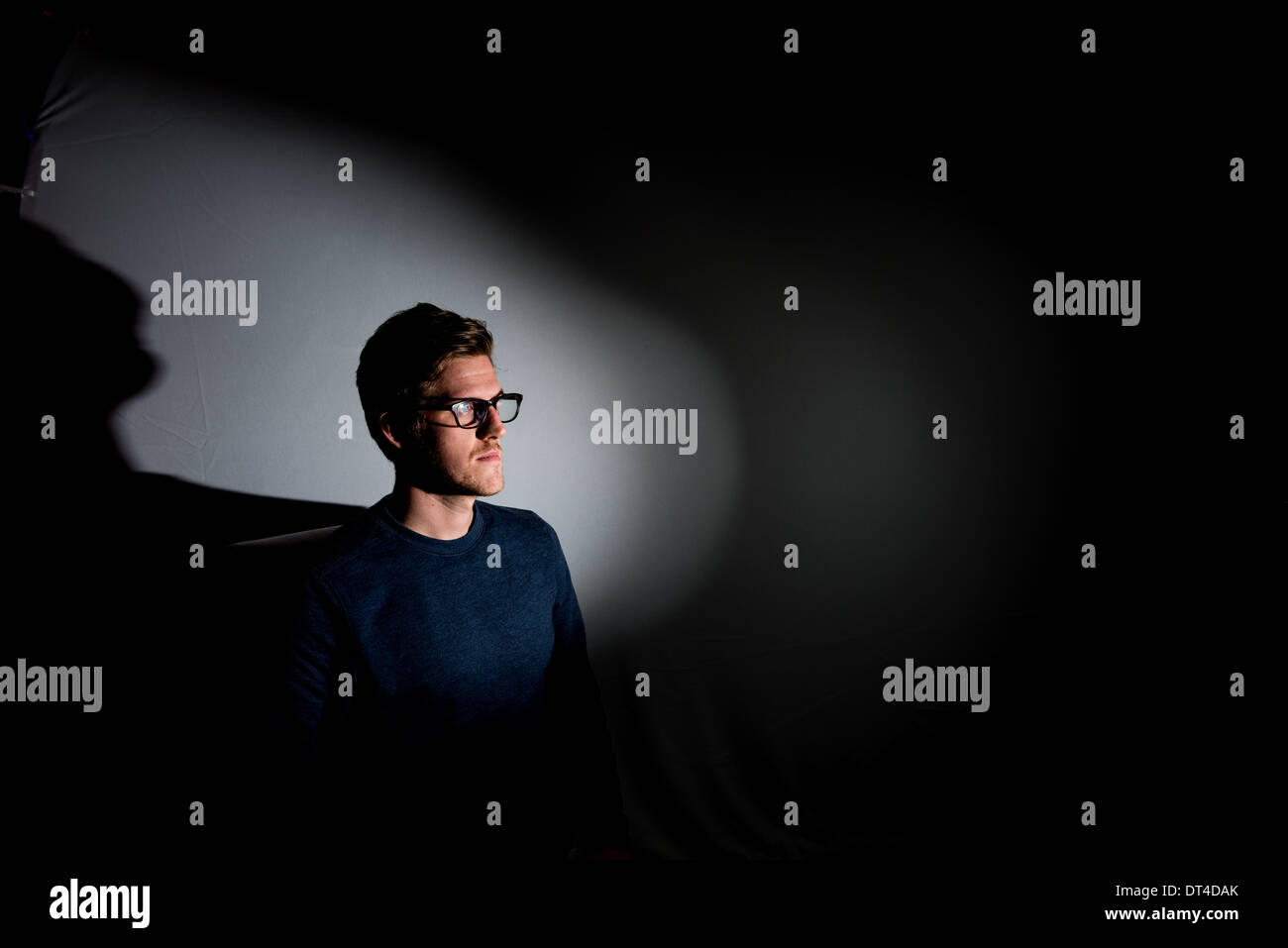 A young man wearing glasses and photographed on black with a shaft of flash light looks away from camera and out of the frame. Stock Photo