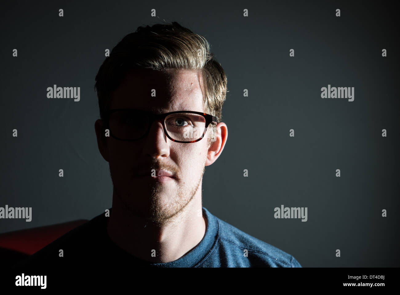 A young man wearing glasses and photographed on black with one side of his face in shadow looks at the camera Stock Photo