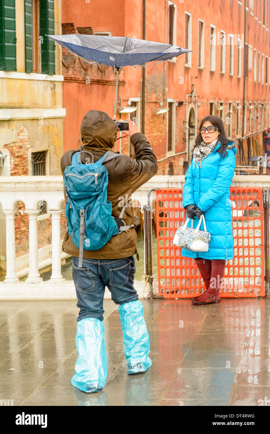 Venice, Italy. Man carrying a inverted umbrella, takes pictures of an Asian women in rainwear on a bridge. Stock Photo