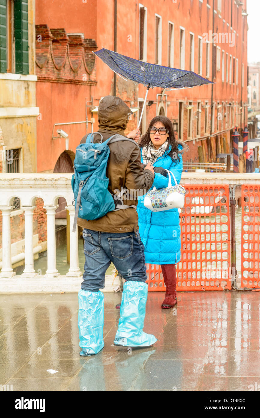 Venice, Italy. Man carrying a inverted umbrella, takes pictures of an Asian women in rainwear on a bridge. Stock Photo