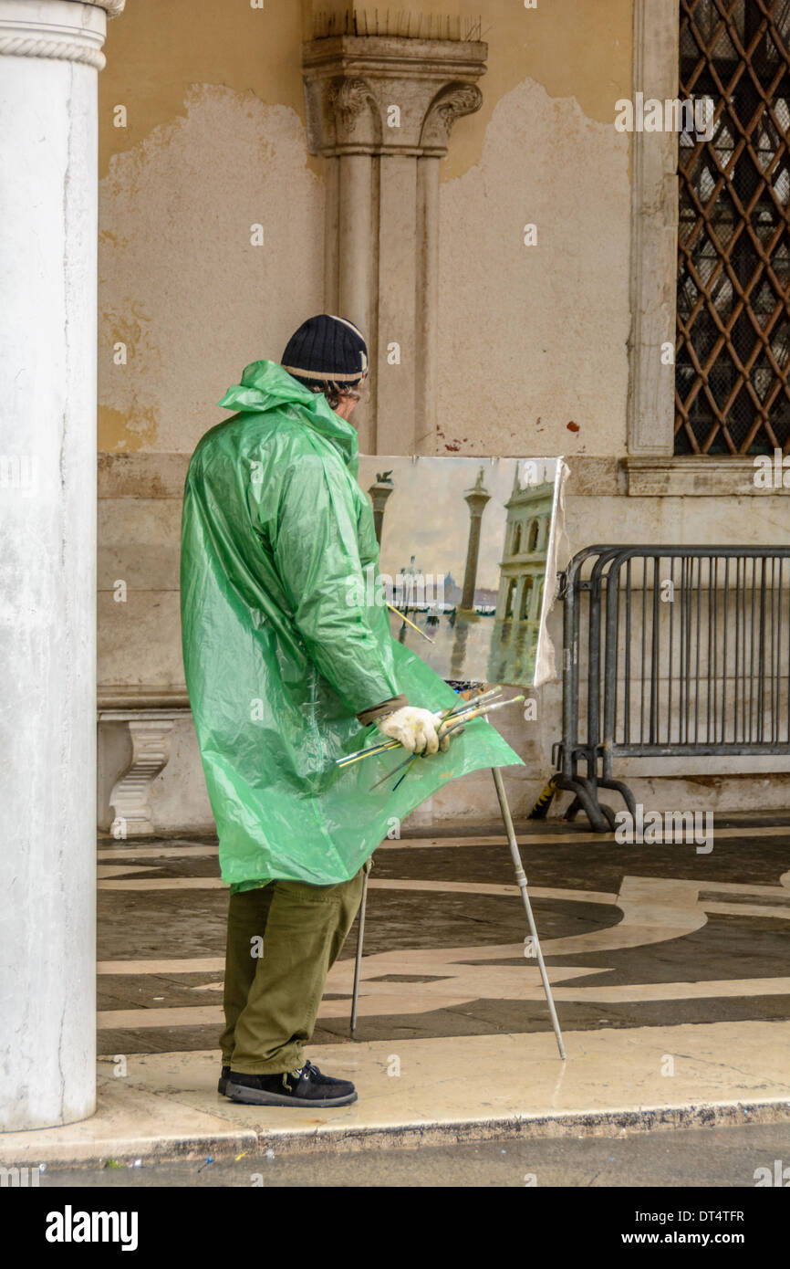 Venice, Italy. Venetian artist with green plastic raincoat paints an oil painting of the Piazzetta San Marco. Stock Photo