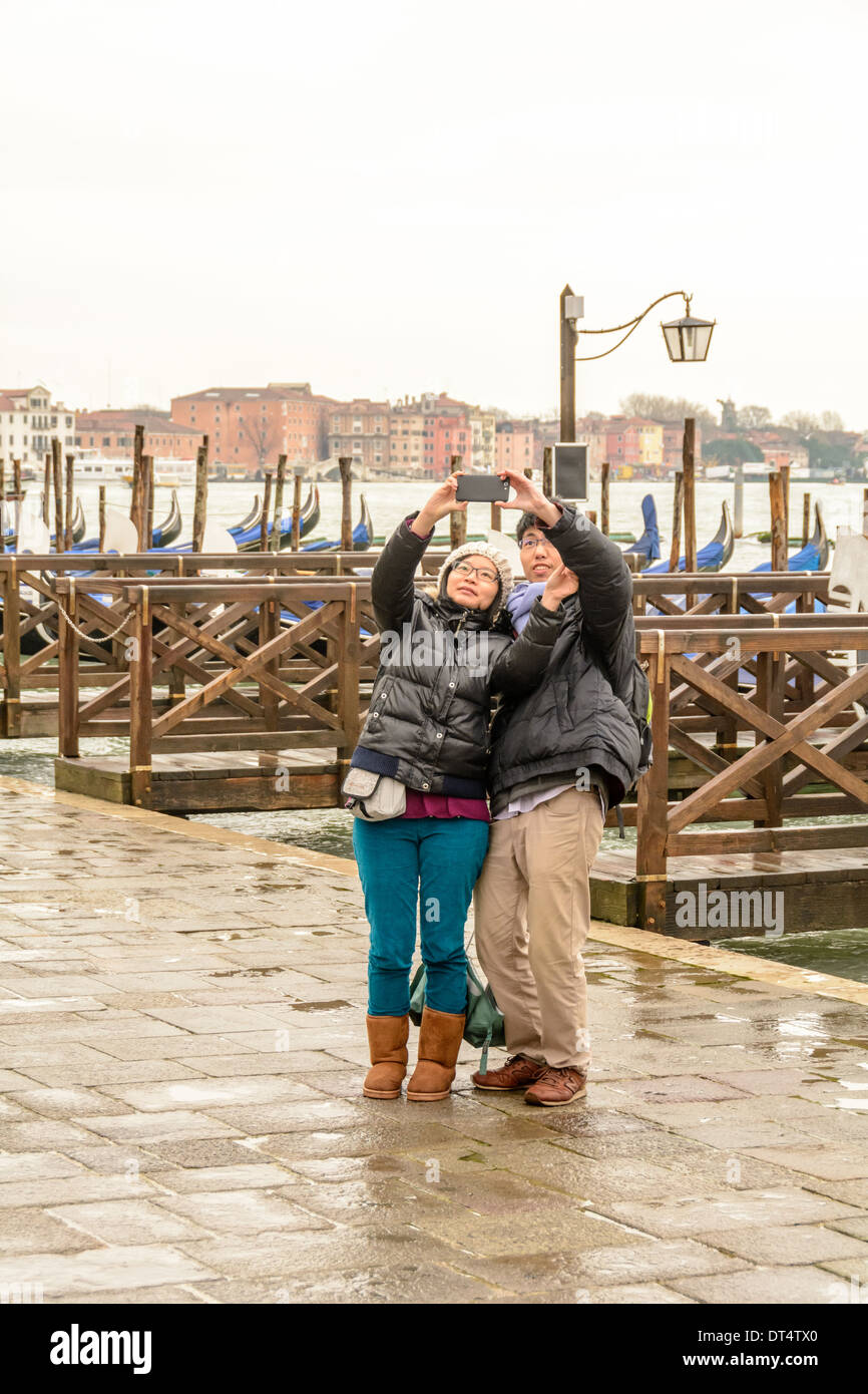Venice, Italy. Youn Asian couple in wet weather clothing standing on the mole with Venetian gondolas in the background taking pictures of themselfs with a smartphone. Stock Photo