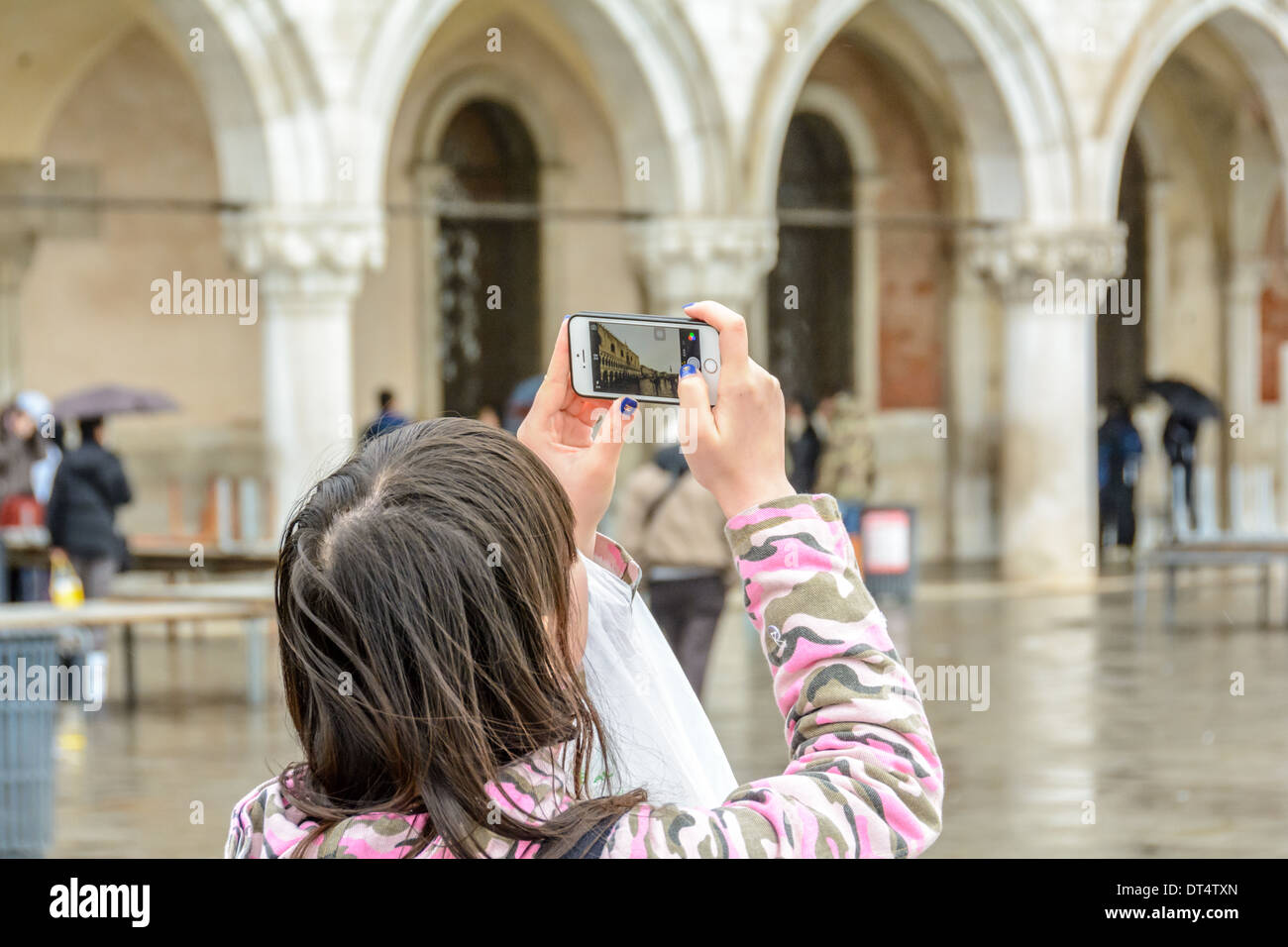 Venice, Italy. Asian girl in pink camouflage dress and blue varnished fingernails with Christian Dior logo, takes pictures of St Mark´s Square with a smartphone. Stock Photo