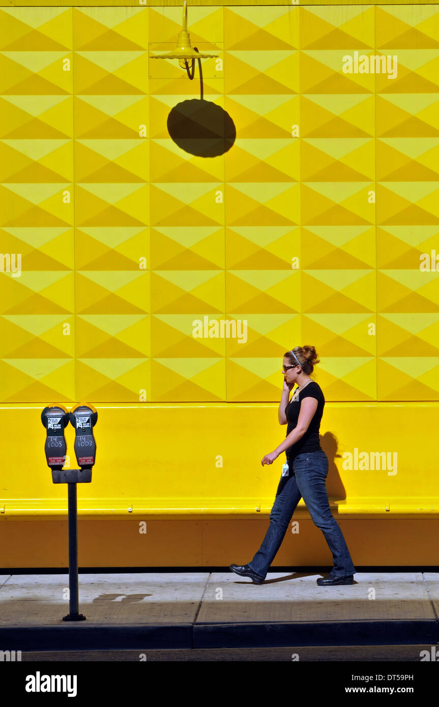 Girl on cell phone walking in front of yellow graphic pattern & shadows on a wall in downtown Denver, Colorado. Stock Photo