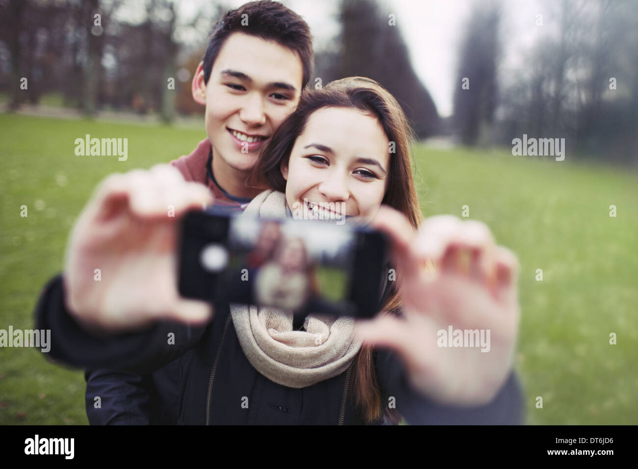 Young couple looking happy while taking pictures using a smart phone at the park. Teenage boy and girl in love photographing. Stock Photo