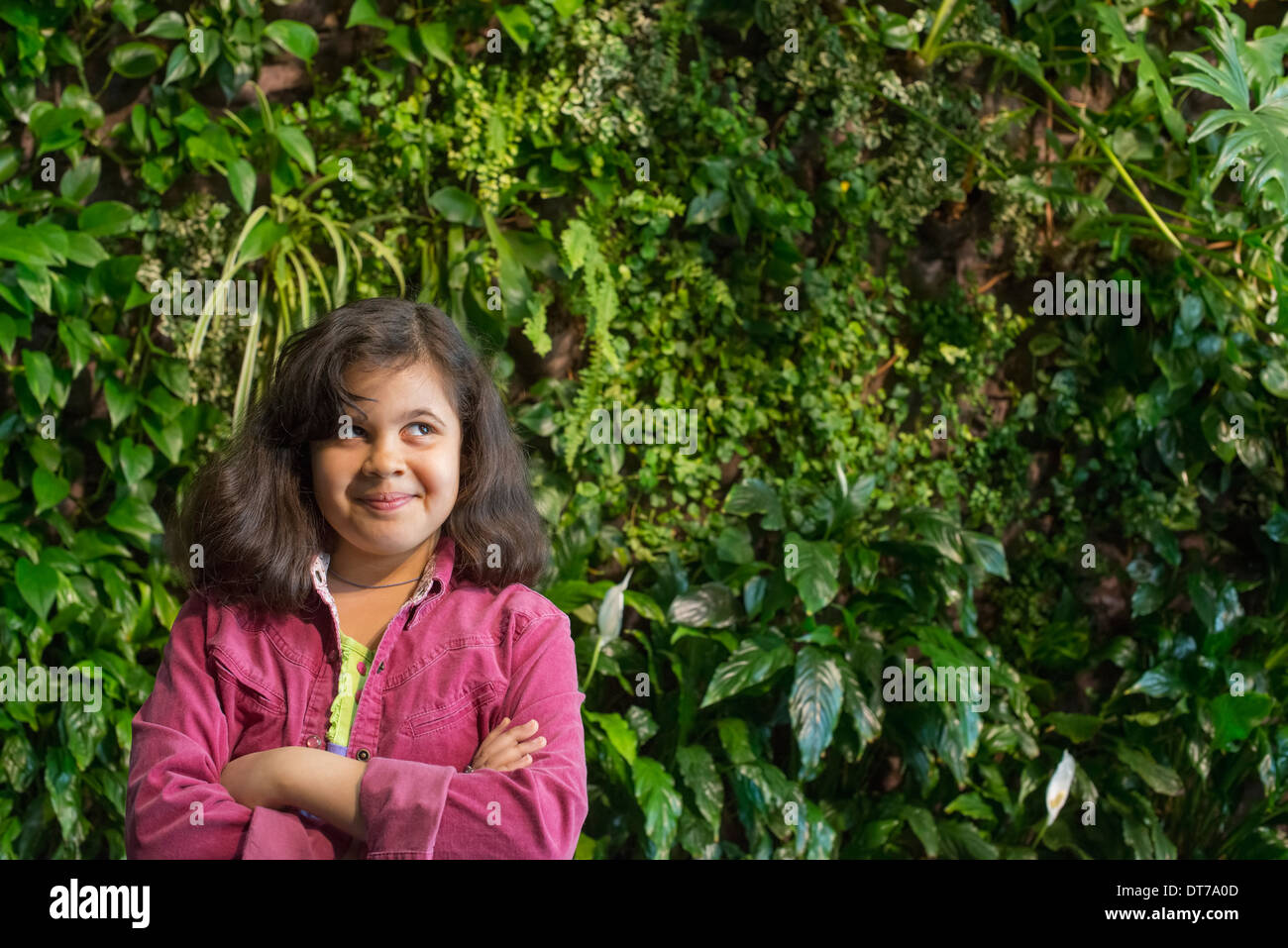 A young girl standing in front of a wall covered with ferns and climbing plants. Stock Photo