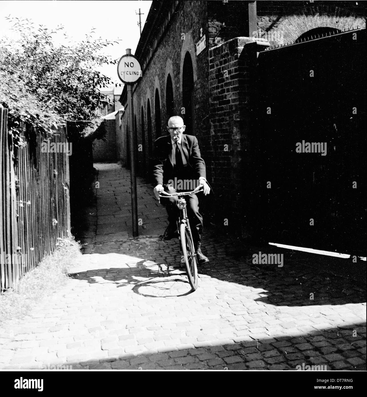 1950s, elderly gentleman in suit on a bicycle coming down a narrow alleyway with sign saying no cycling. Stock Photo