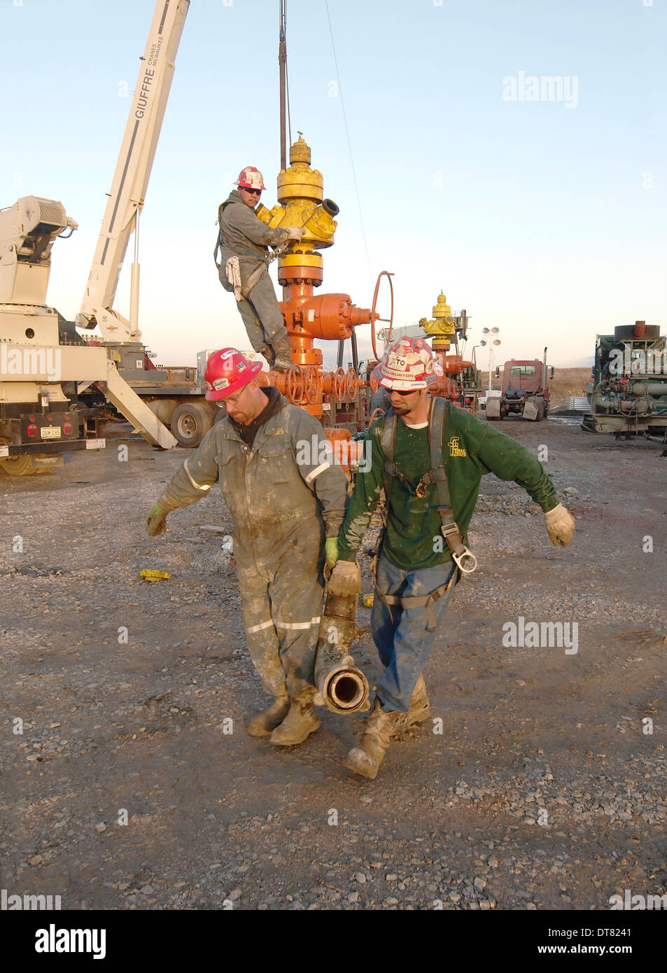 Two workers to carry a heavy fitting to attach pipe to well-head as they prepare to frack the well. Stock Photo