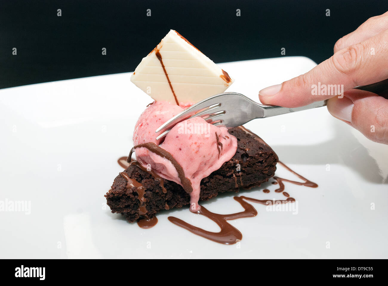 Close-up of a man's hand using a fork to get a taste of brownie with melting raspberry ice-cream and a white chocolate garnish. Stock Photo