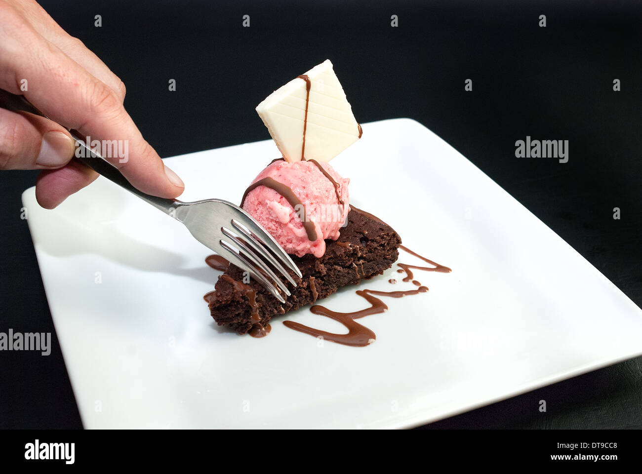Close-up of a man's hand using a fork to get a taste of brownie with raspberry ice-cream and a white chocolate garnish. Stock Photo