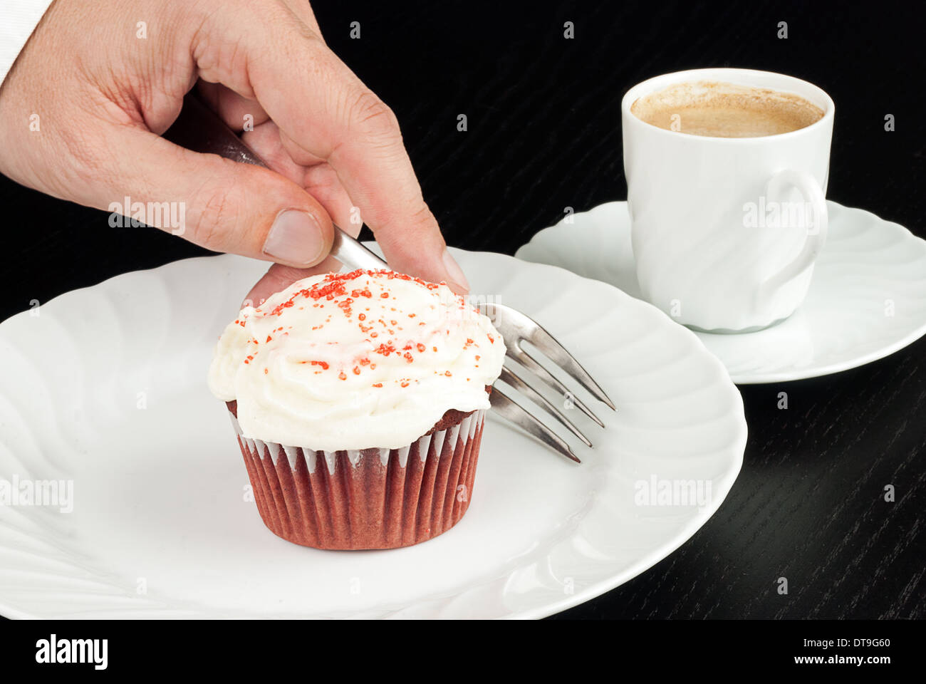 Close-up of a red velvet cupcake on a plate with a man's hand picking up a fork to eat it, espresso in the background. Stock Photo