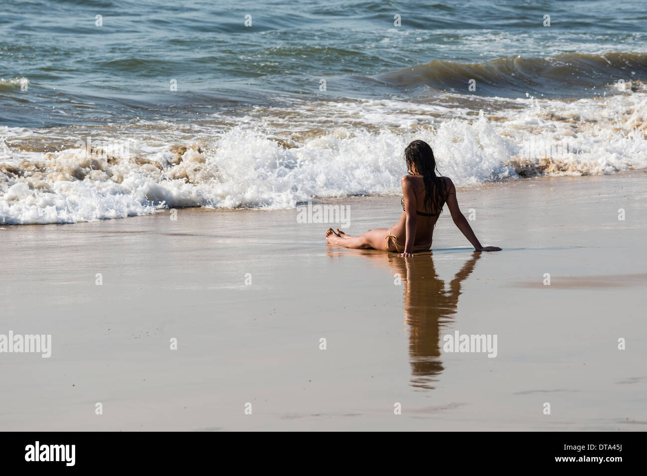 A young woman wearing bikini is sitting in the sand of Anjuna Beach, Anjuna, Goa, India Stock Photo
