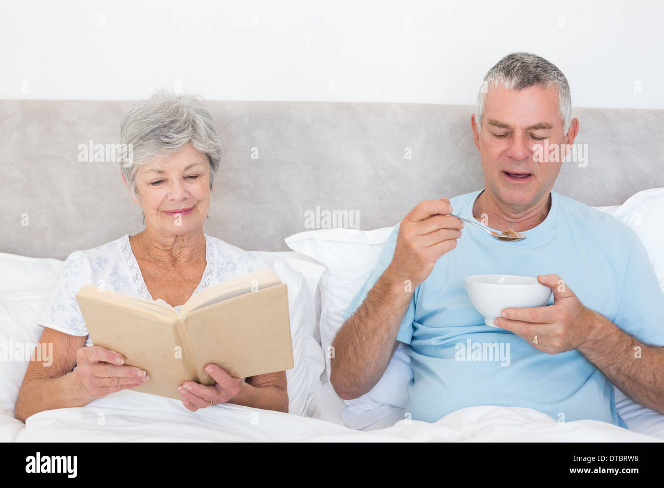Senior couple with book and cereal bowl in bed Stock Photo