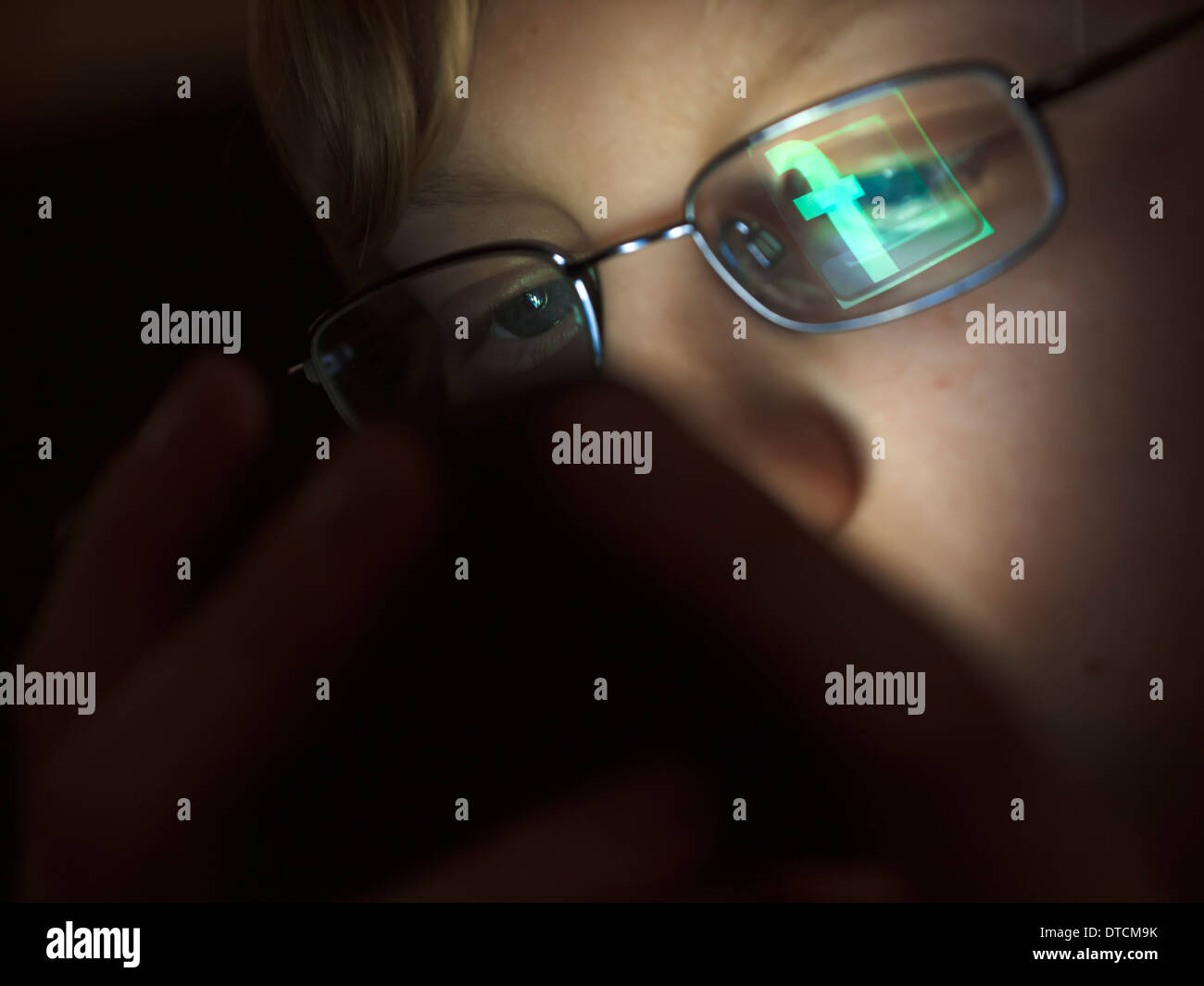 A teenage boy is surfing the internet using his iPhone, with the Facebook logo being reflected in the boy's glasses. Stock Photo