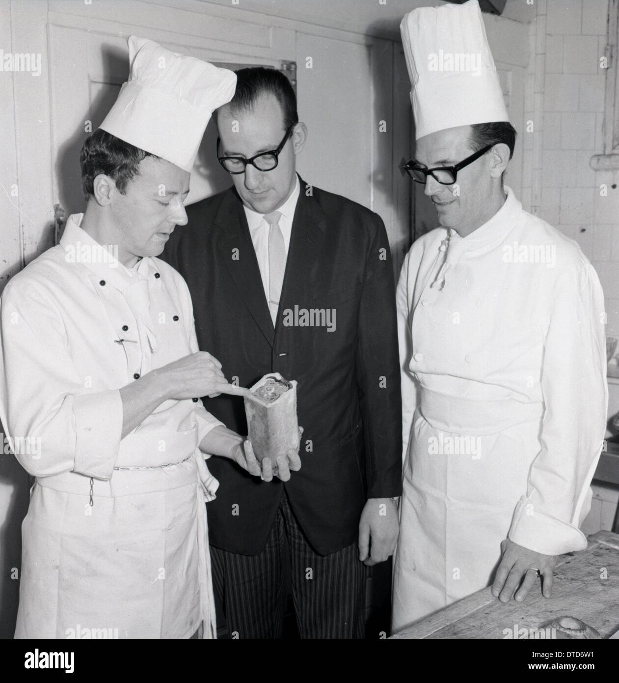1960s. Historical image showing two professional chefs in a kitchen in conversation with a hotel maitre d' about a food item. Stock Photo