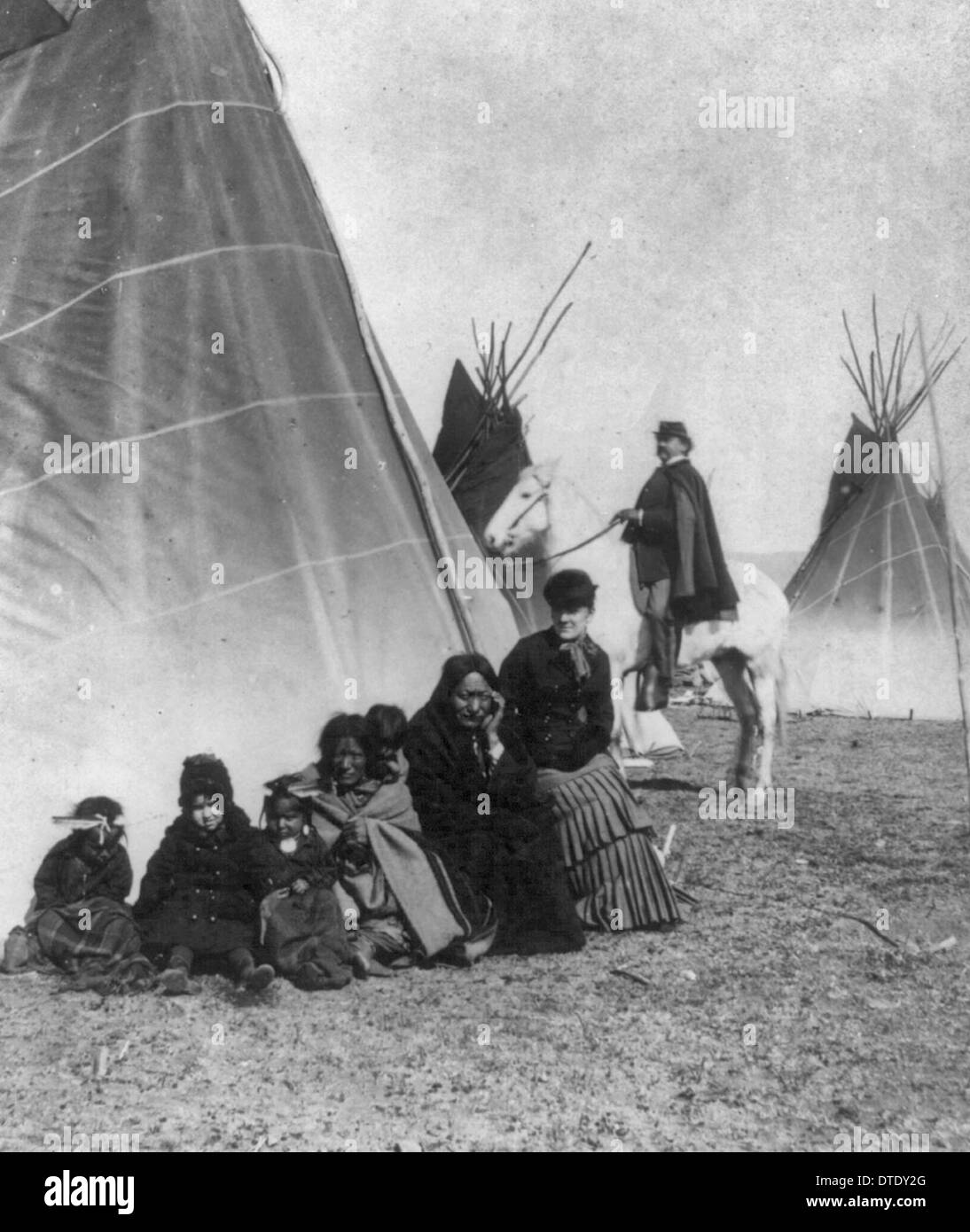 Sitting Bull, squaw and twins - Seated in front of tepee with a White woman and child; papoose on his squaw's back; Cavalry officer on horseback in background. 1882 Stock Photo