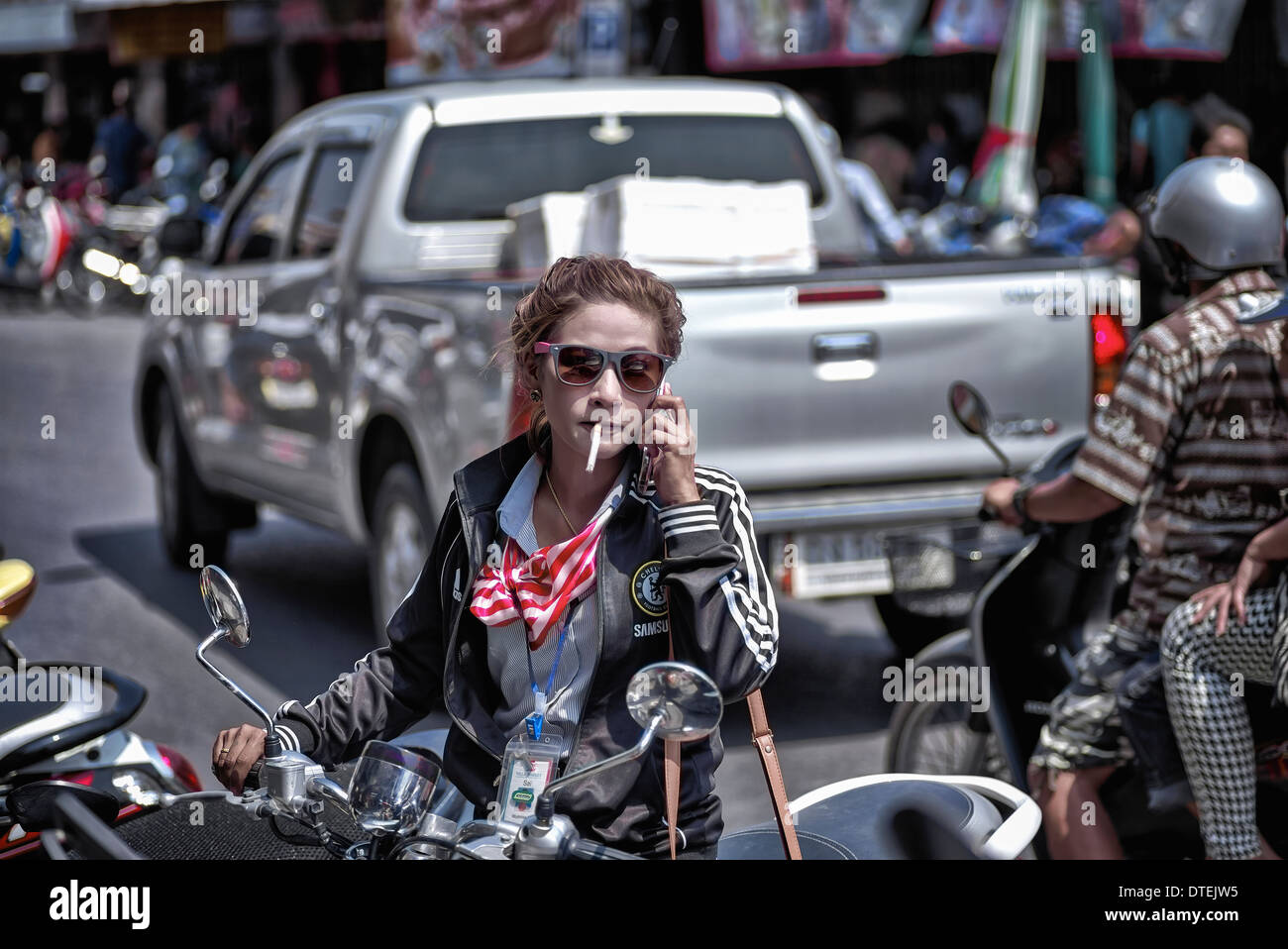 Woman smoking a cigarette and using a mobile phone whilst preparing to ride her motorcycle. Thailand S. E. Asia Stock Photo