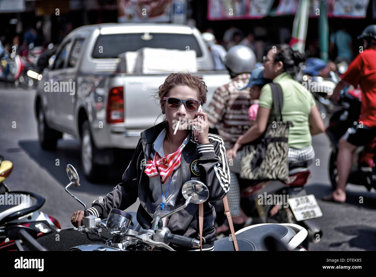 Woman smoking a cigarette and using a mobile phone whilst preparing to ride her motorcycle. Thailand S. E. Asia Stock Photo