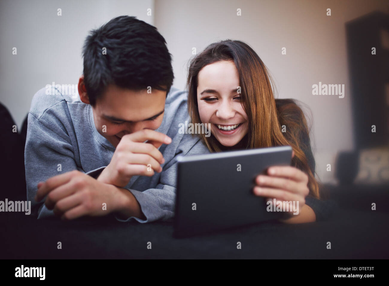 Happy young man and woman using tablet PC. Mixed race teenage couple using digital tablet smiling while lying on sofa at home. Stock Photo