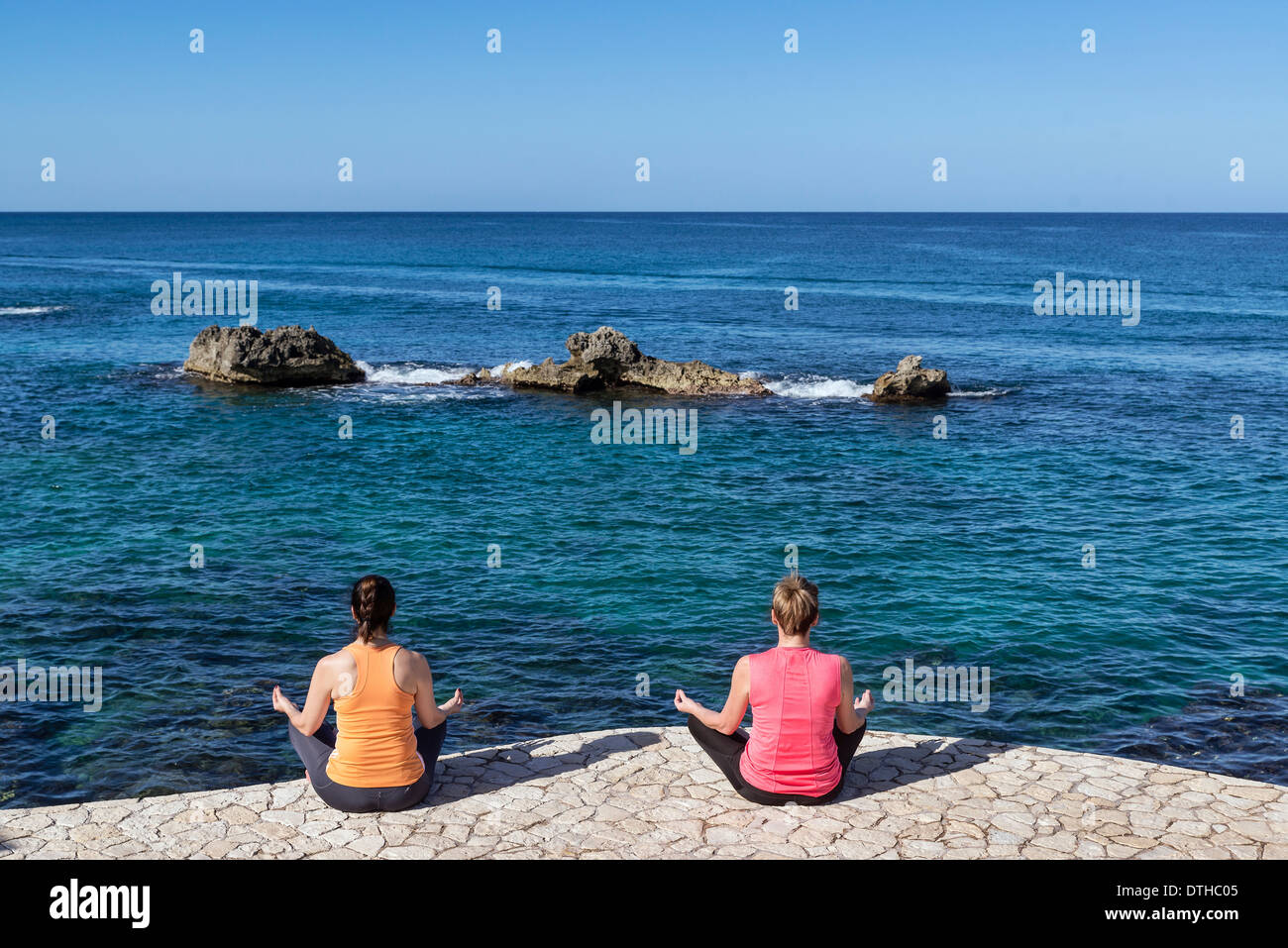 Women practice yoga meditation from an ocean waterfront ledge, Negril, Jamaica. Stock Photo