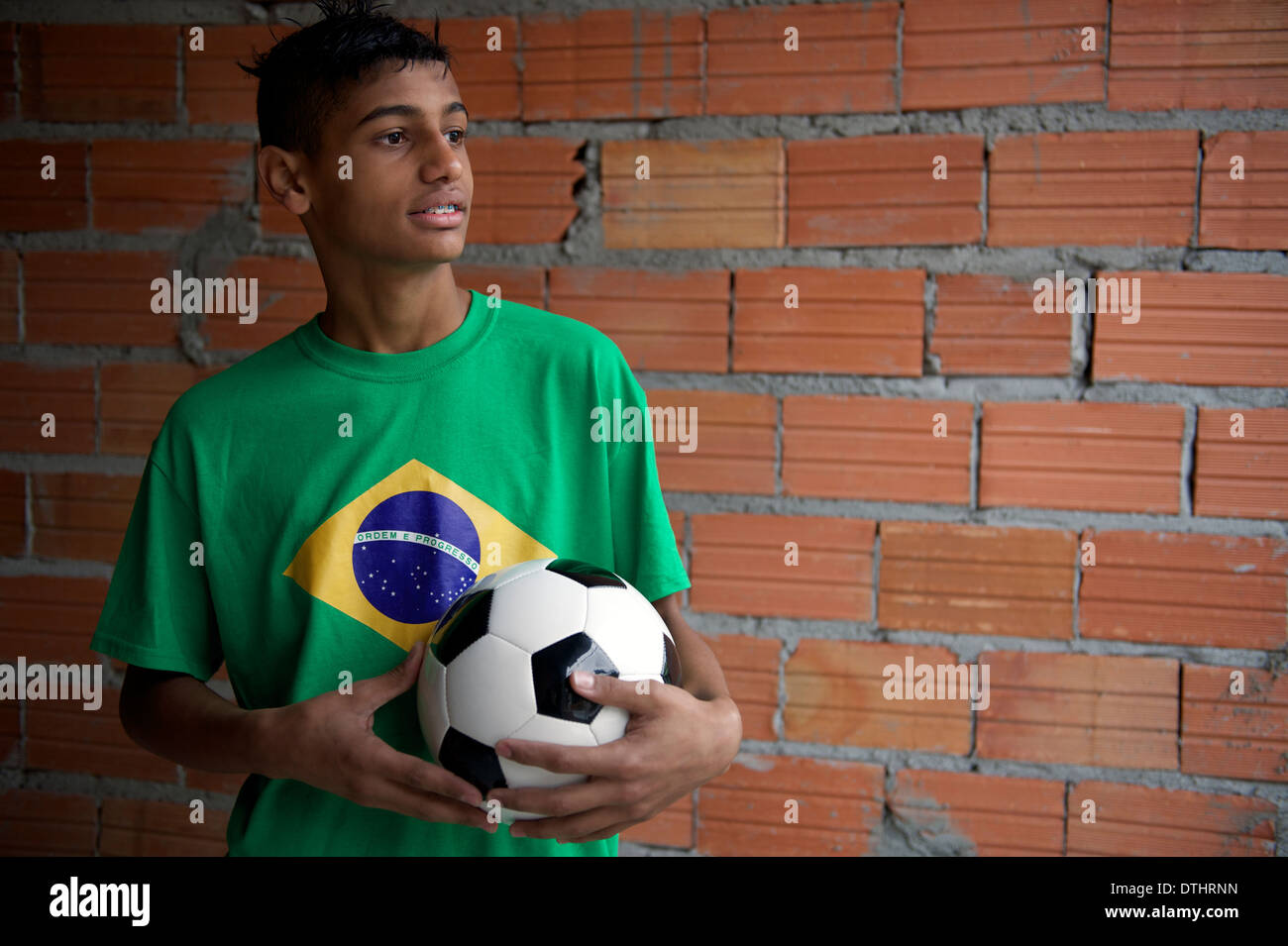 Portrait of Brazilian teen wearing Brazilian flag t-shirt holding football soccer ball near simple favela house wall Stock Photo