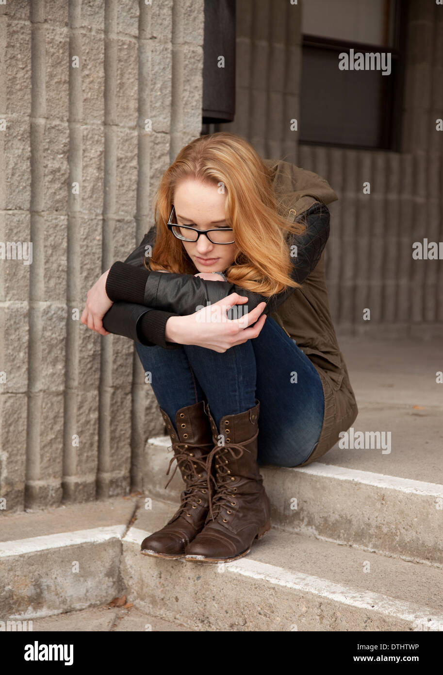 Outdoor photo of young teenage girl seated on concrete steps, hugging knees, distraught facial expression. Stock Photo