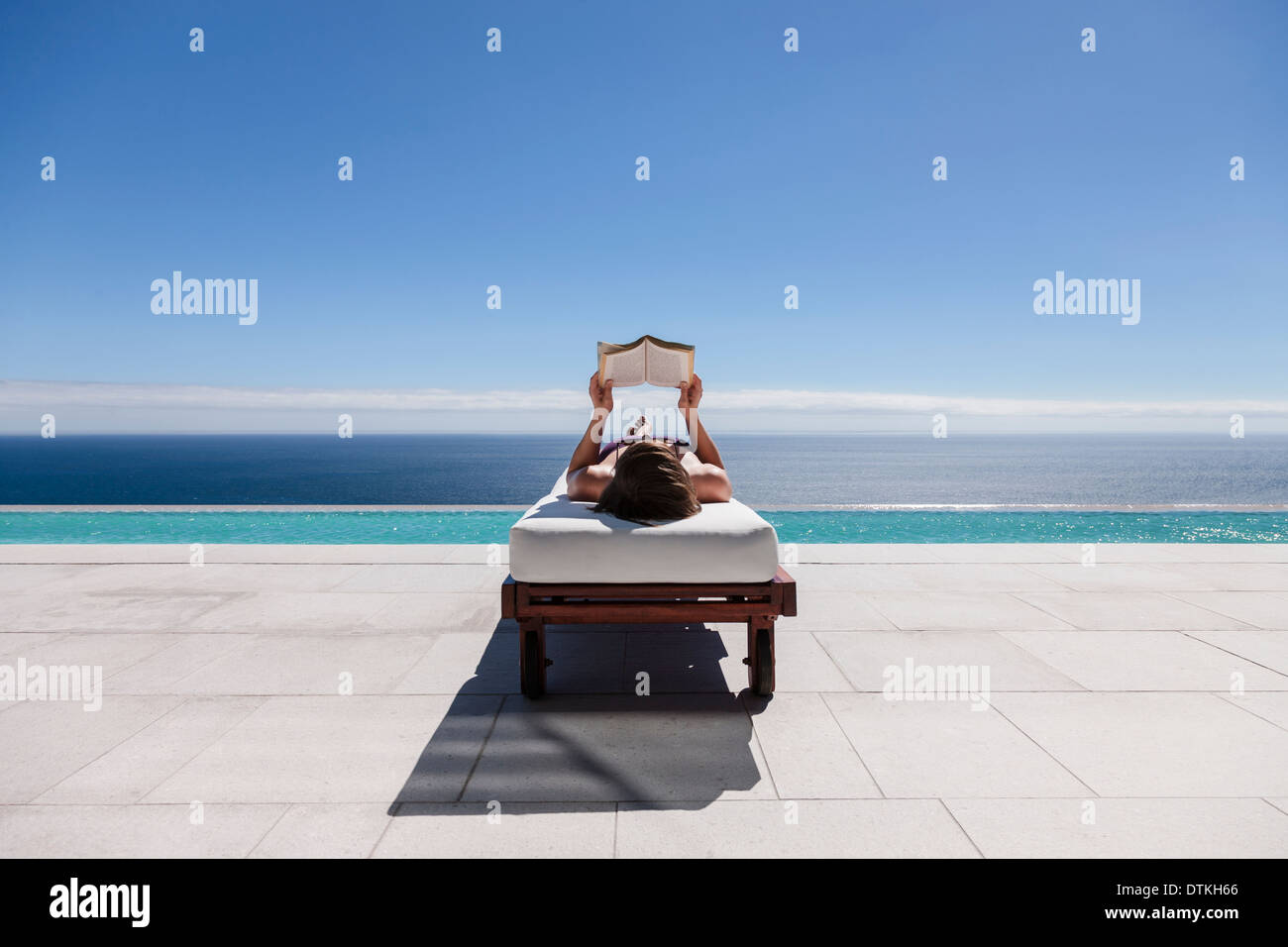 Woman Reading On Lounge Chair At Poolside Overlooking Ocean Stock