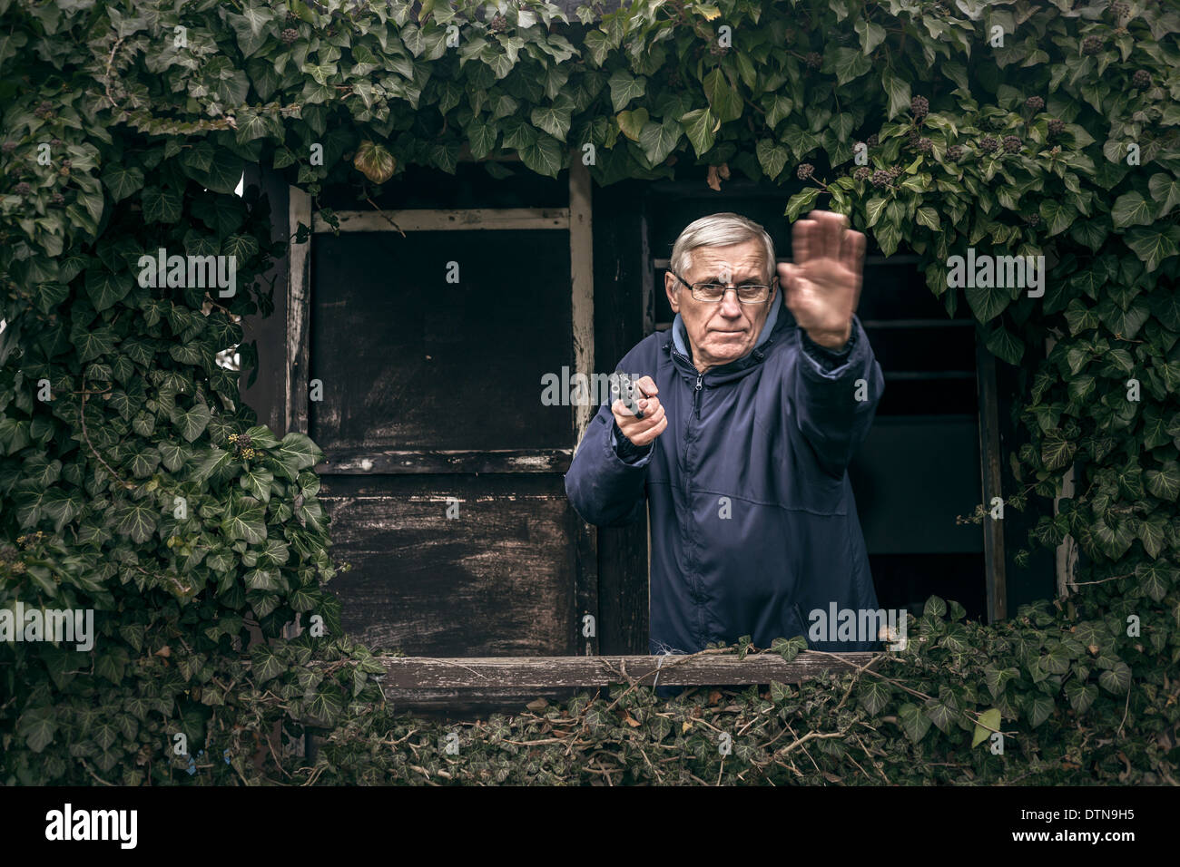 Furious senior man gesturing with a gun protecting his property, standing in front of old overgrown cabin. Stock Photo