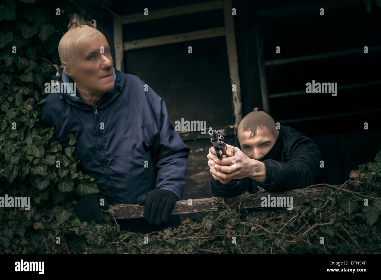 Two masked armed men hiding on overgrown porch of old cabin. Stock Photo