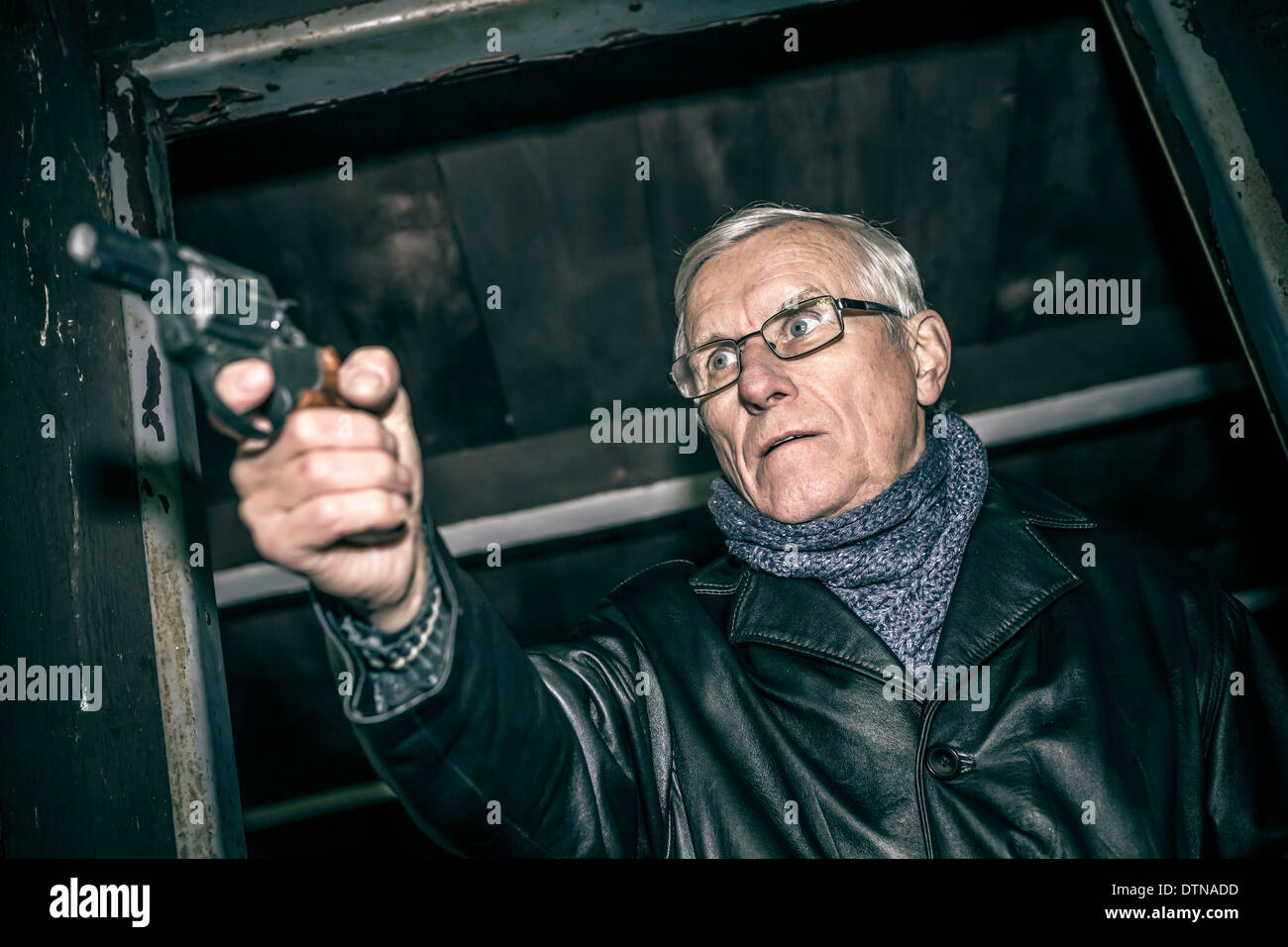 Dangerous senior man aiming a gun and standing in old dark cabin. Stock Photo