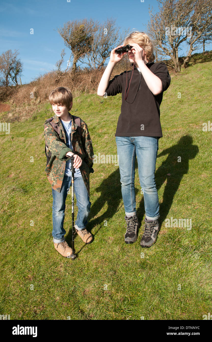 Two teenage boys standing in a field, one is looking through a pair of binoculars and the other holds a walking stick Stock Photo