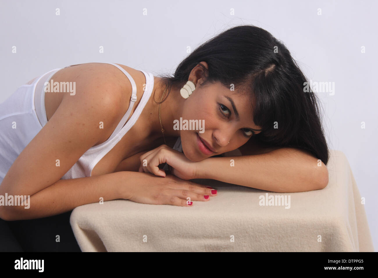 Beautiful young, black-haired Latino in a white singlet, leaning over a small table, resting her head on the back of her arms Stock Photo