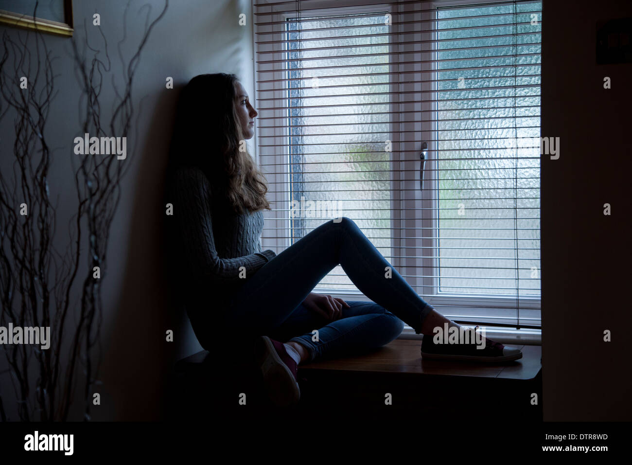 Young female with long hair wearing jeans, sad and looking towards a window, sitting in darkness at home. Side viewpoint. Stock Photo