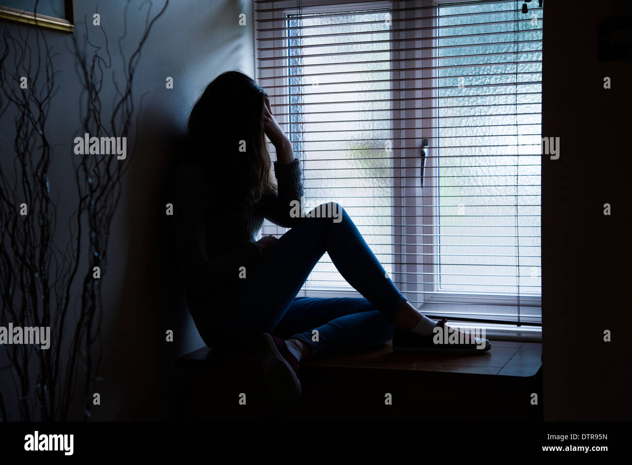 Girl with long hair wearing jeans, hand on head in despair, sitting in darkness by a window at home. Side viewpoint. Stock Photo
