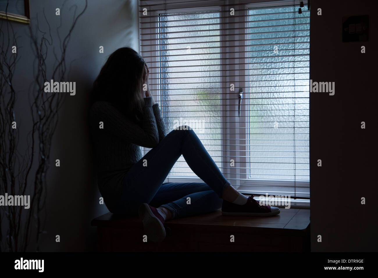Girl with long hair wearing jeans, hands covering, sitting in darkness by a window at home. Side viewpoint. Stock Photo