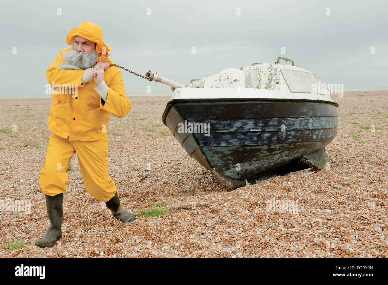 Old fisherman in yellow water-proof overalls, pulling an old fishing boat along the pebble beach. Stock Photo