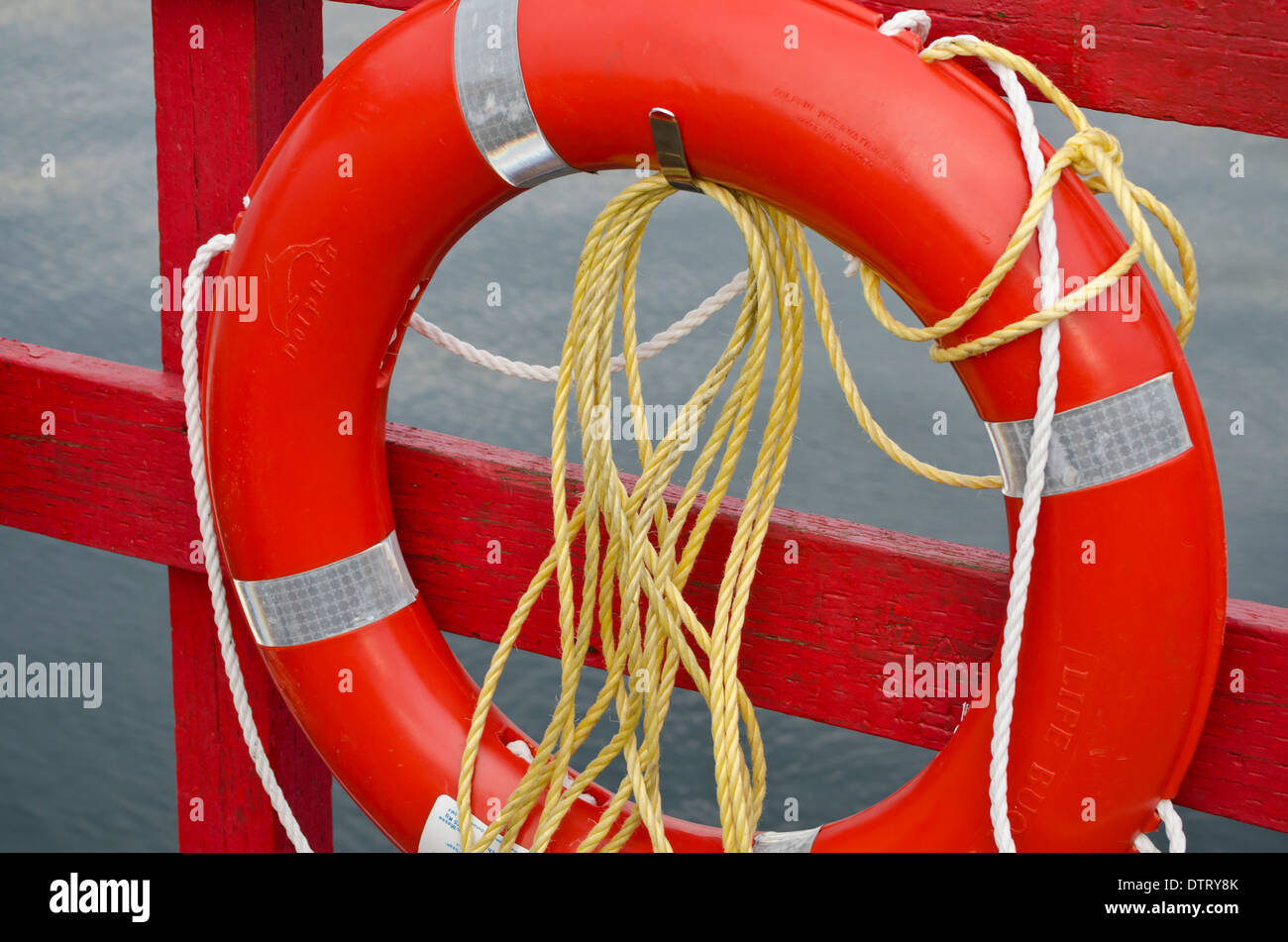 Red round life preserver and yellow ropes tied against a red post by the water. Stock Photo