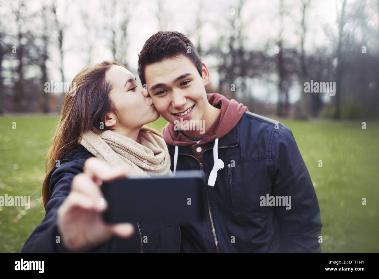 Pretty young girl kissing her boyfriend on cheeks while taking self portrait with a mobile phone. Mixed race couple in park. Stock Photo