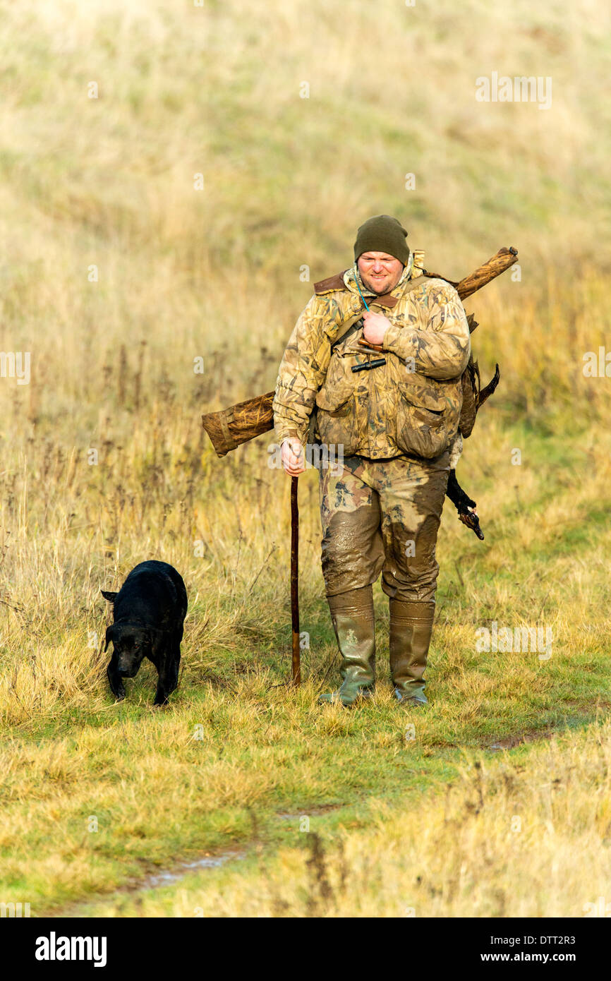Wildfowler walking over salt marsh with dog and huge canada goose over his shoulder. 1 of 5 images taken. Stock Photo