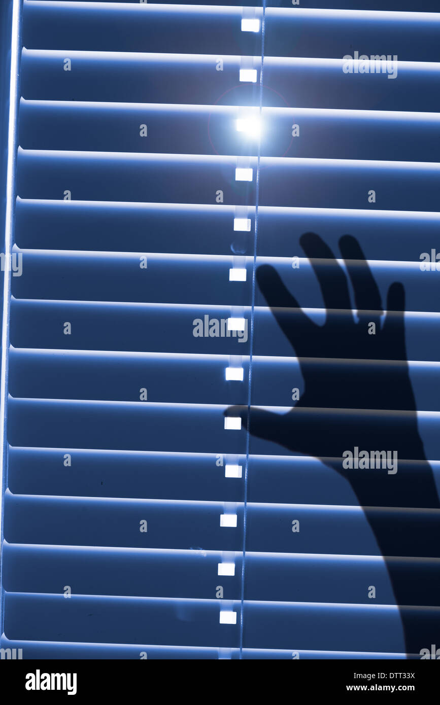 Conceptual image of being trapped and helpless. Shadow of a hand reaching for closed window blinds with sunlight outside. Stock Photo