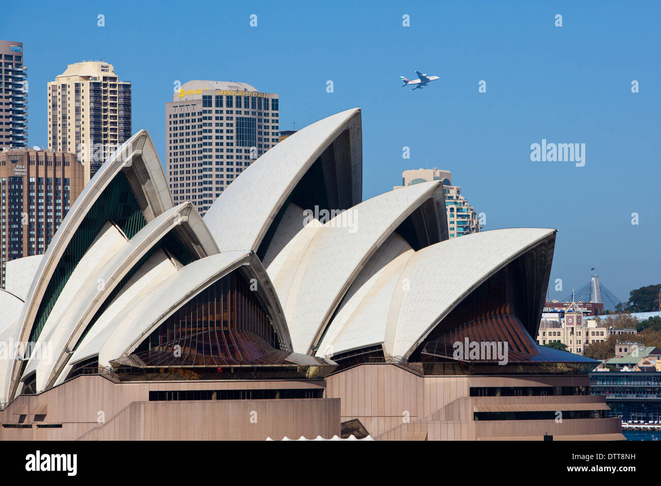Sydney Opera House from the harbour with waterfront buildings beyond and airliner taking off from airport flying overhead Stock Photo