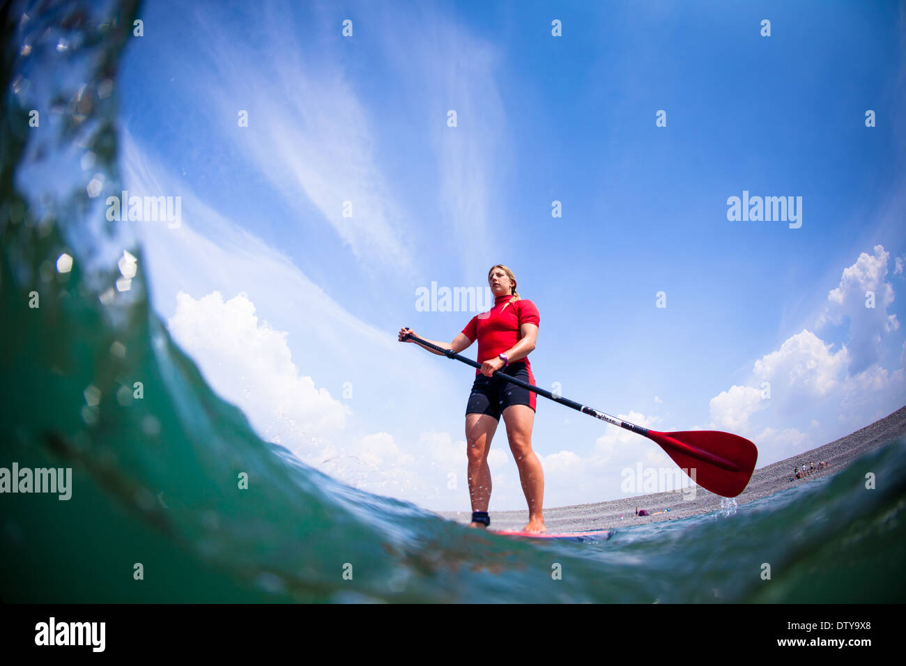 A girl in a red rash vest paddleboards in the clear waters of North Devon UK SUP (stand up paddleboarding) Stock Photo
