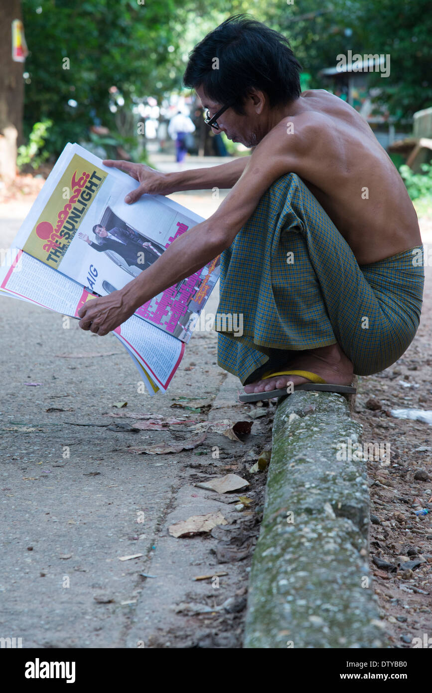Man reading a newspapaer in the street while squatting on a pavement. Yangon. Myanmar (Burma). Stock Photo