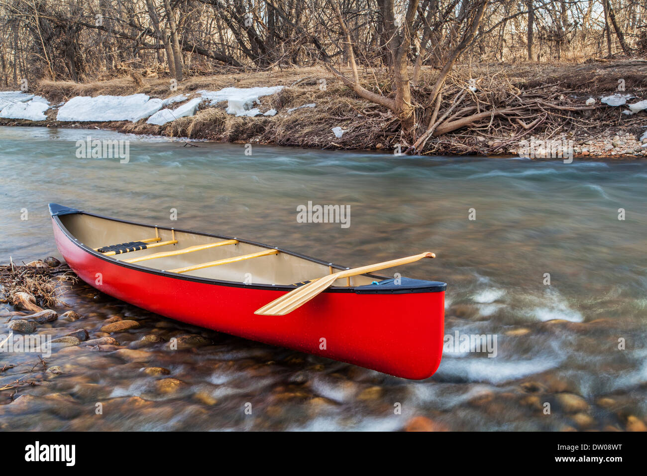 red canoe with a wooden paddle on river shore in winter or early spring - Cache la Poudre River, Fort Collins, Clorado Stock Photo