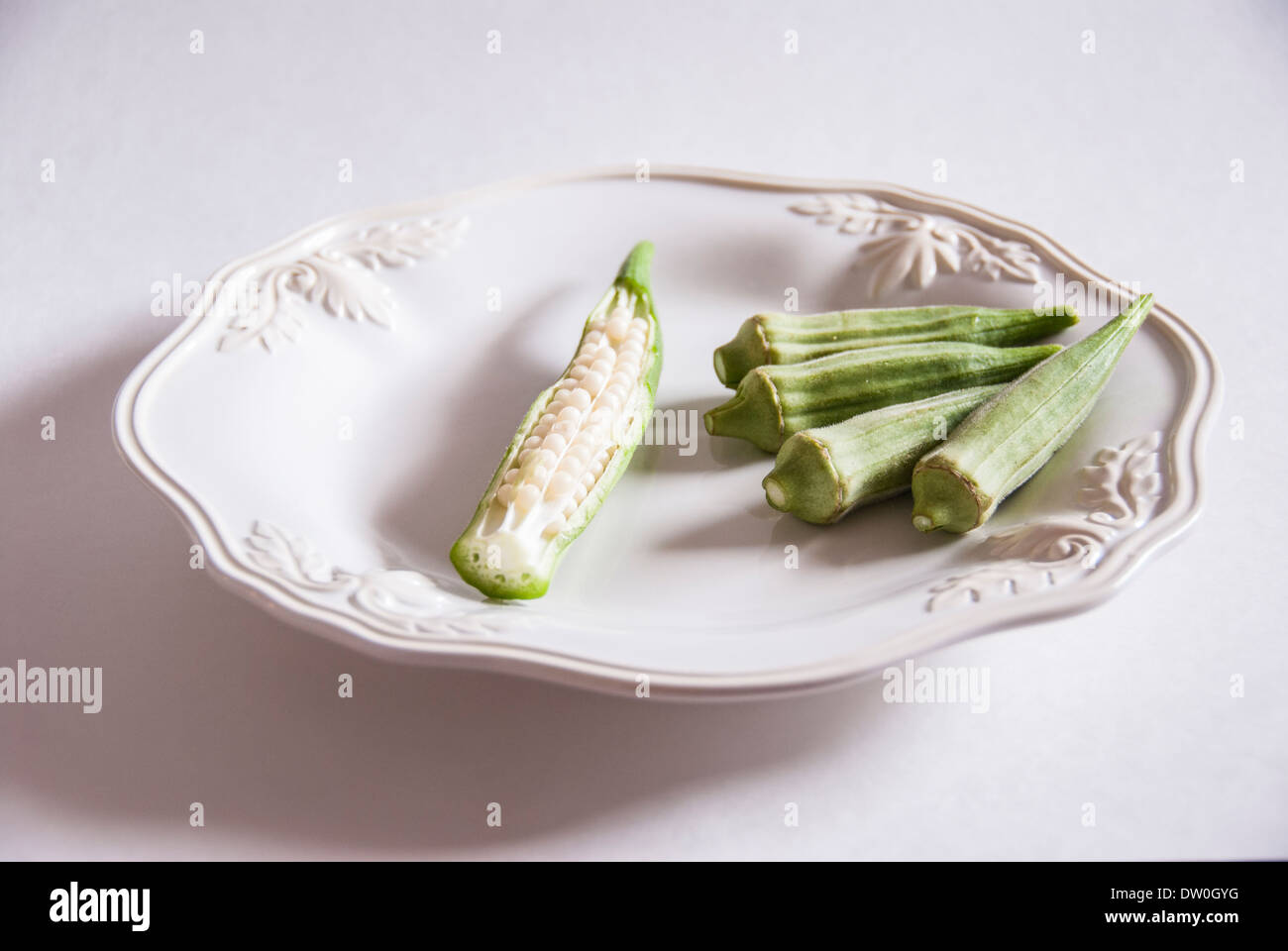 Fresh uncooked okra pods on a white dish, with one cut open showing seeds. Stock Photo