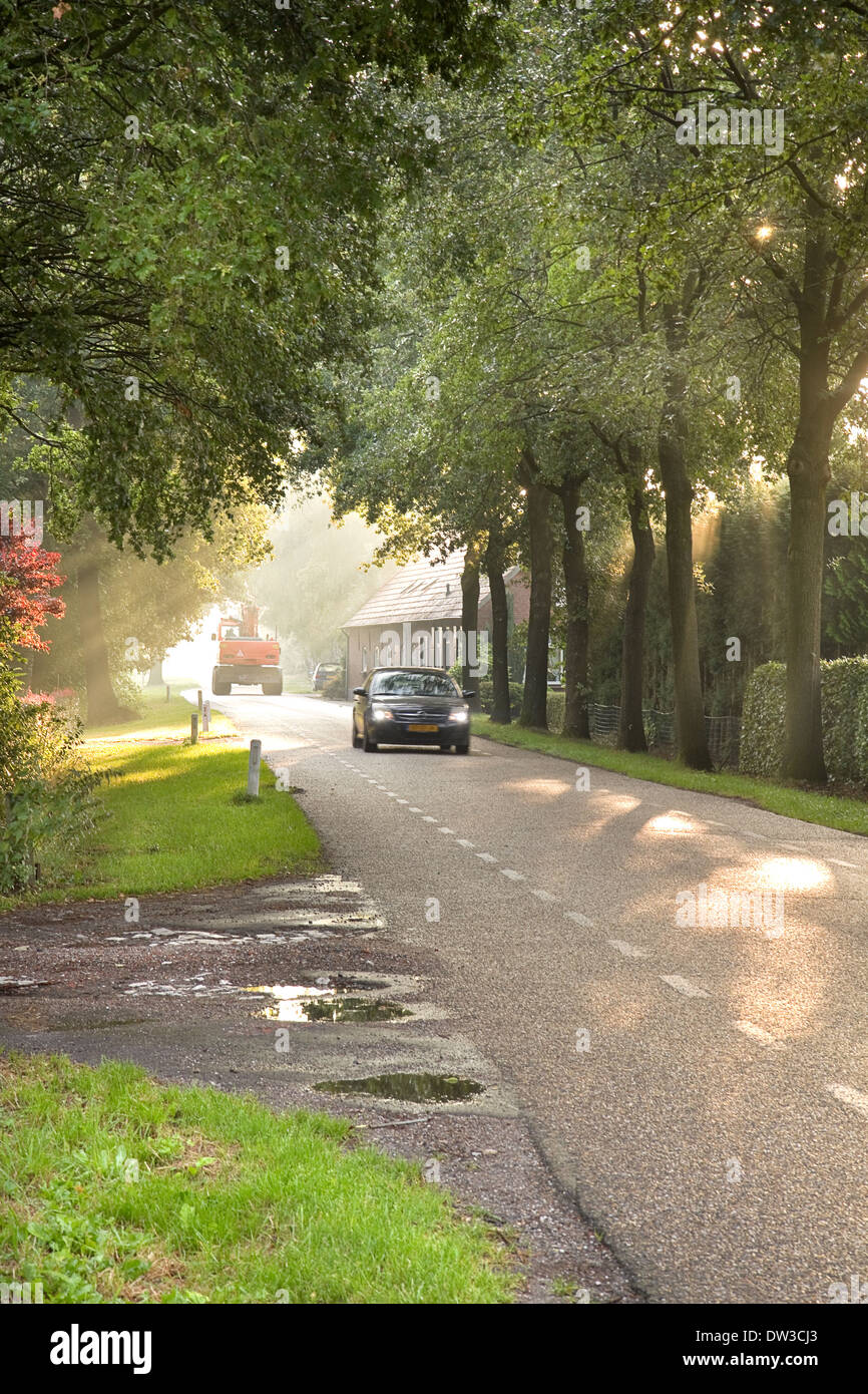 Country road with traffic, farm, trees and sunbeams at sunrise Stock Photo