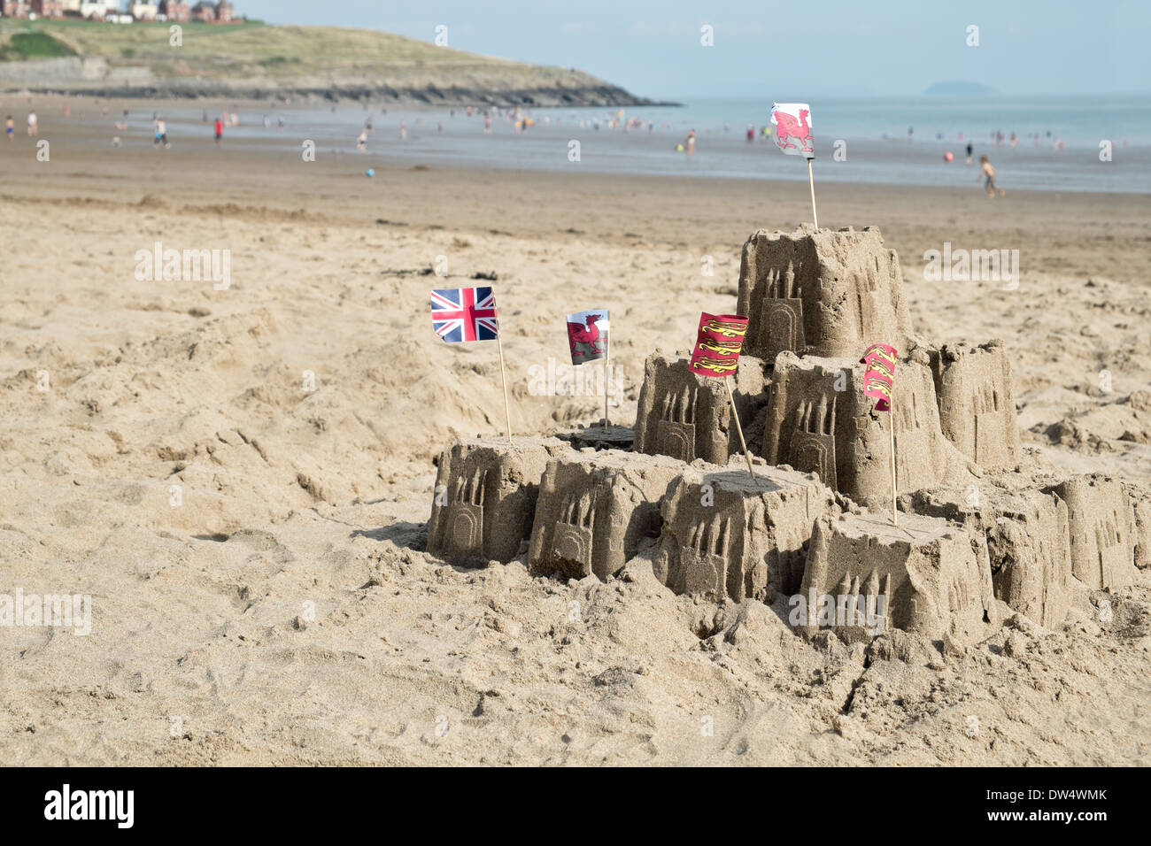 A sandcastle on a welsh beach with the sea behind & holiday makers enjoying the water Stock Photo